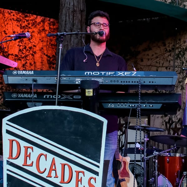 A musician with glasses and a beard sings into a microphone while playing a Yamaha keyboard.