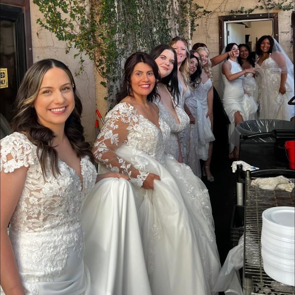 A group of women in white dresses, possibly wedding gowns, smiling and posing together outdoors.