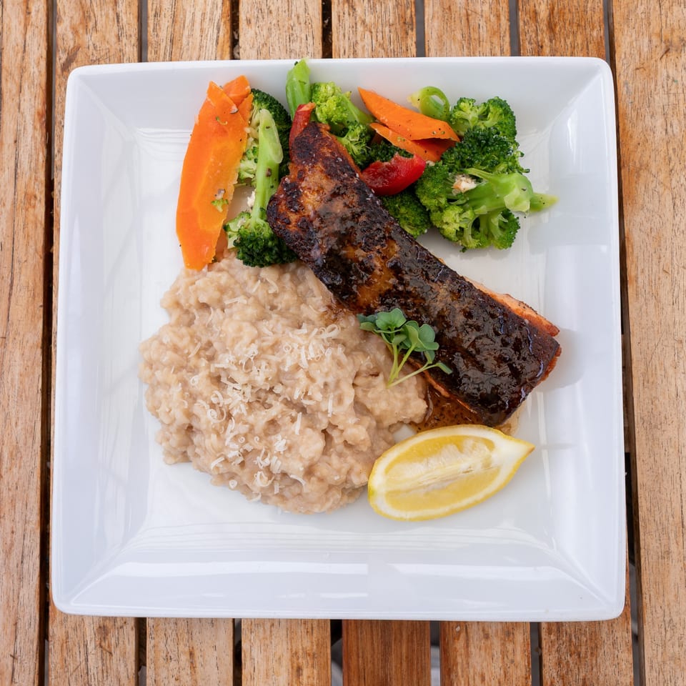 Glazed salmon with risotto, broccoli, carrots, and a lemon wedge on a white plate.
