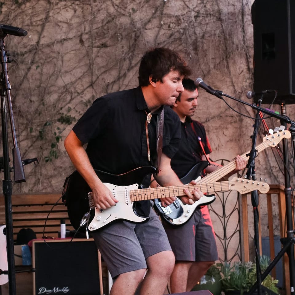 Two men playing electric guitars on a stage with a vine-covered wall behind them.