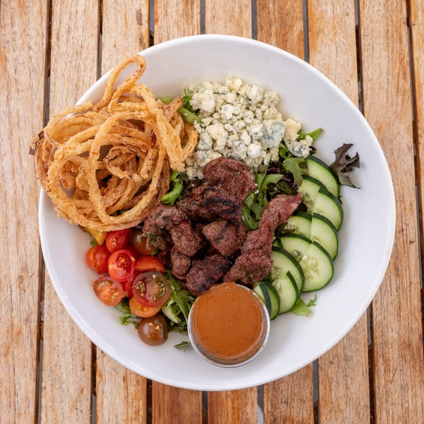 Steak salad with crispy onions, blue cheese, tomatoes, cucumbers, and dressing on a wooden table.