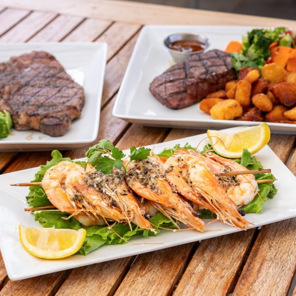 Grilled shrimp skewers with lemon and parsley, and two steak dinners with sides on a wooden table.