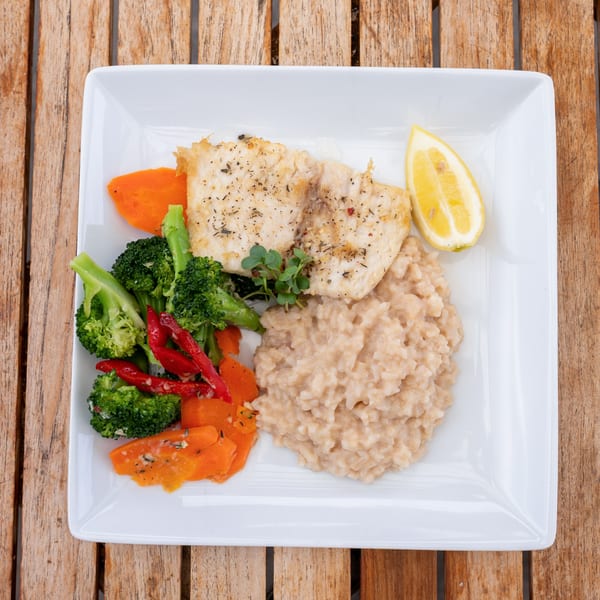 A white plate with grilled fish, risotto, broccoli, carrots, red peppers, and a lemon wedge.