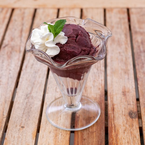 A scoop of dark berry sorbet with whipped cream and a mint leaf in a glass dish on a wooden table.