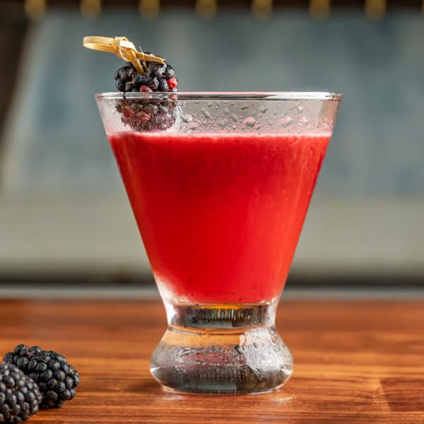 A vibrant red cocktail in a martini glass, garnished with fresh blackberries on a wooden skewer.