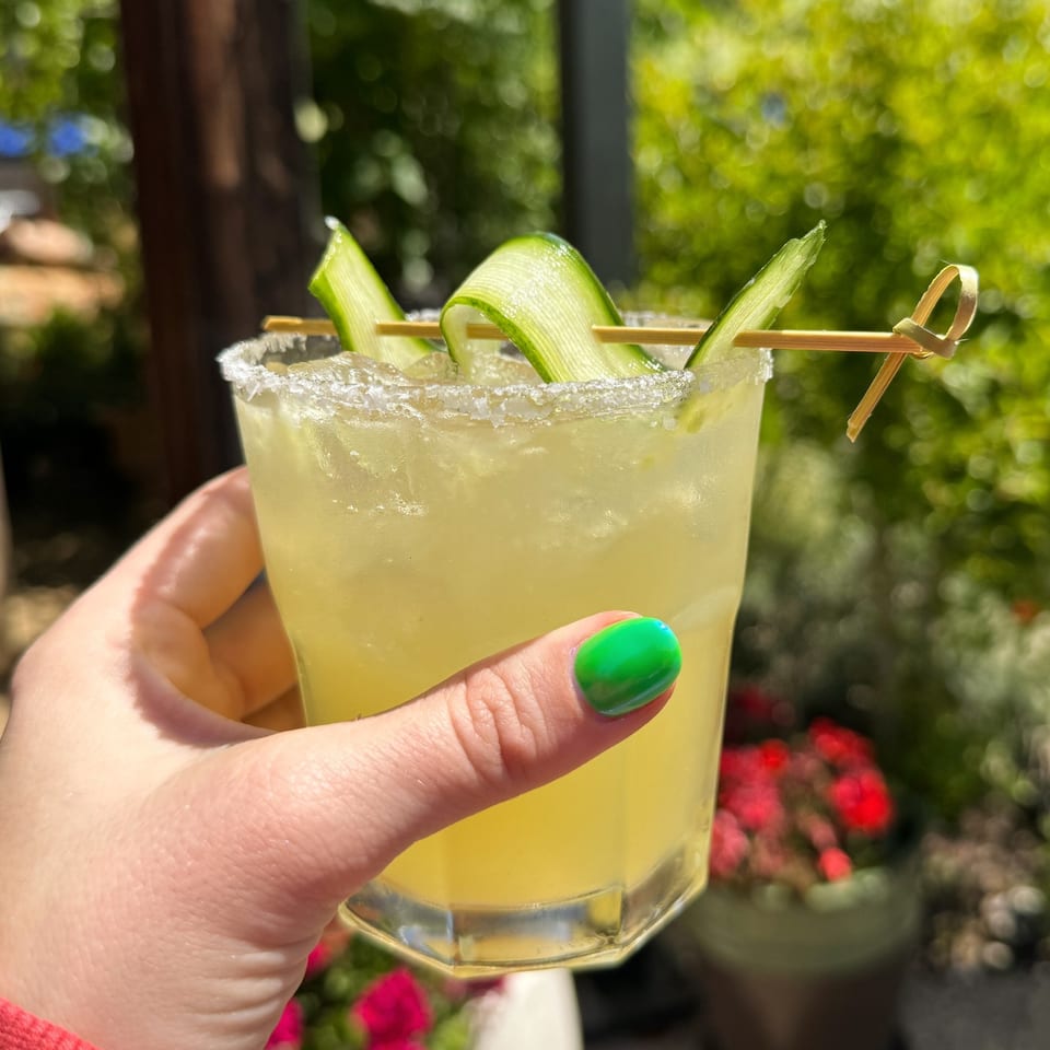 A hand with green nail polish holds a Union cucumber margarita with a salted rim outdoors.
