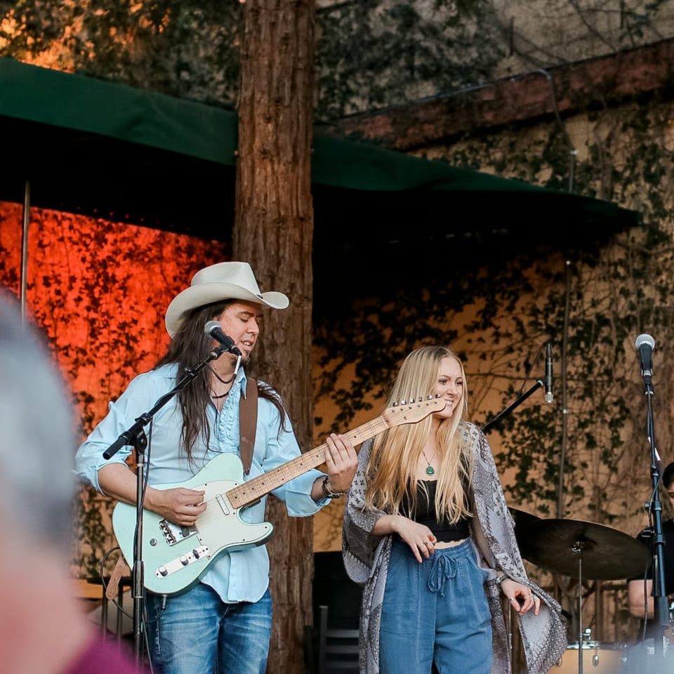 A male guitarist in a cowboy hat and a female singer performing live on an outdoor stage.