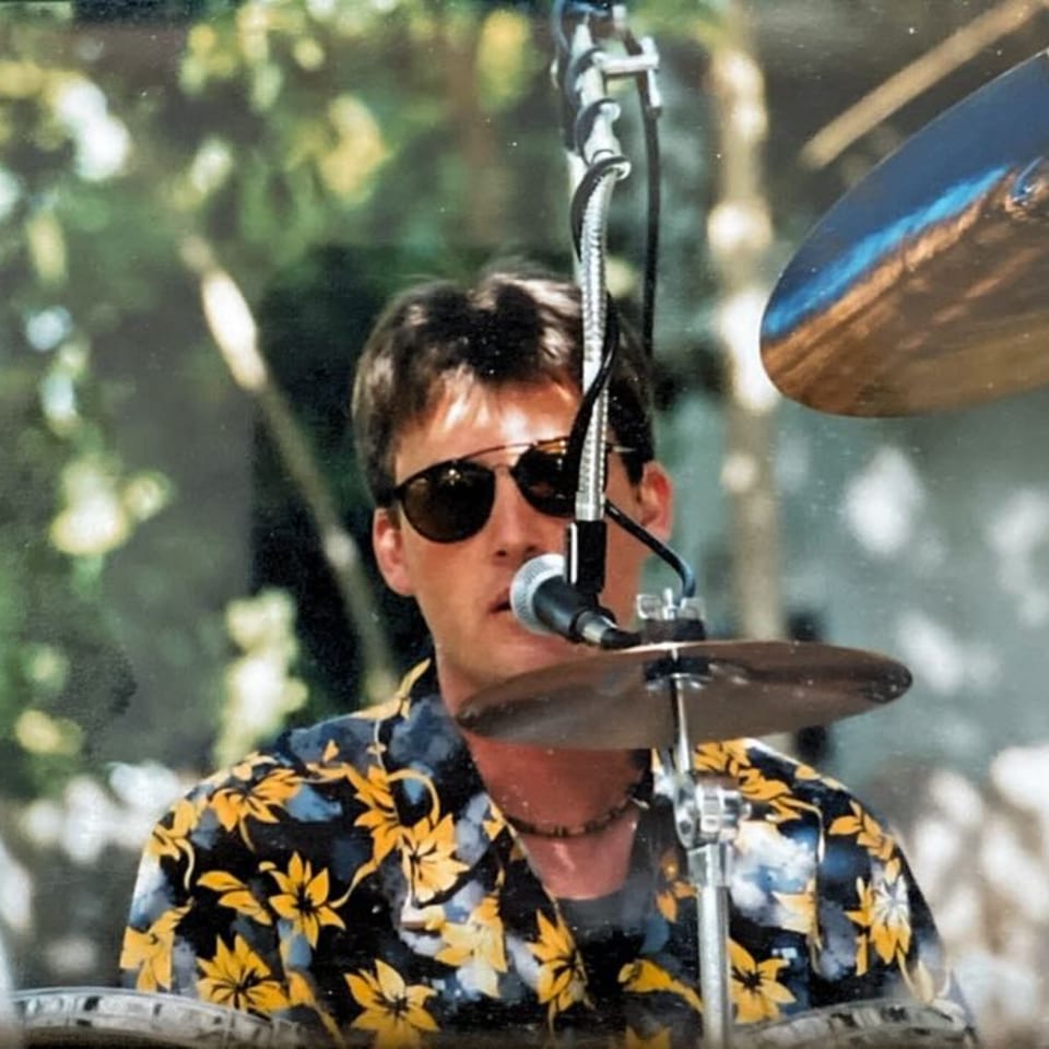 A man in sunglasses and a floral shirt playing drums with a microphone in front of him.