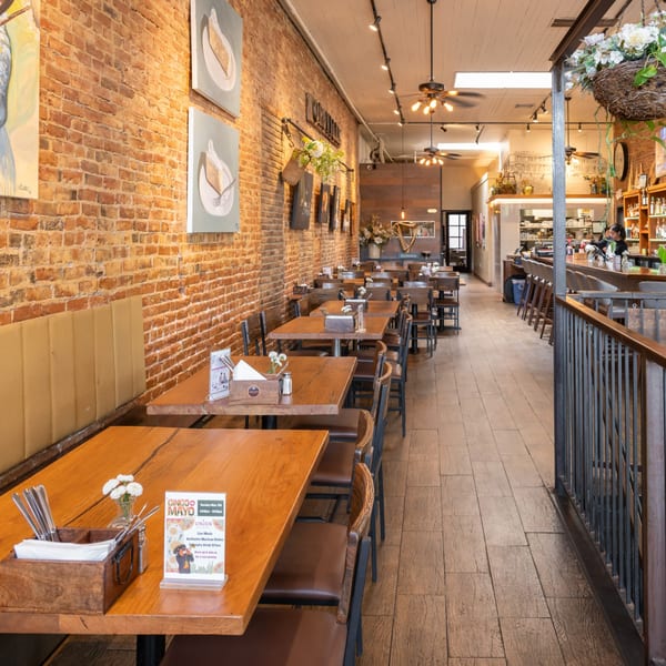Long, narrow restaurant dining room with wooden tables and chairs, brick walls, and a bar area.