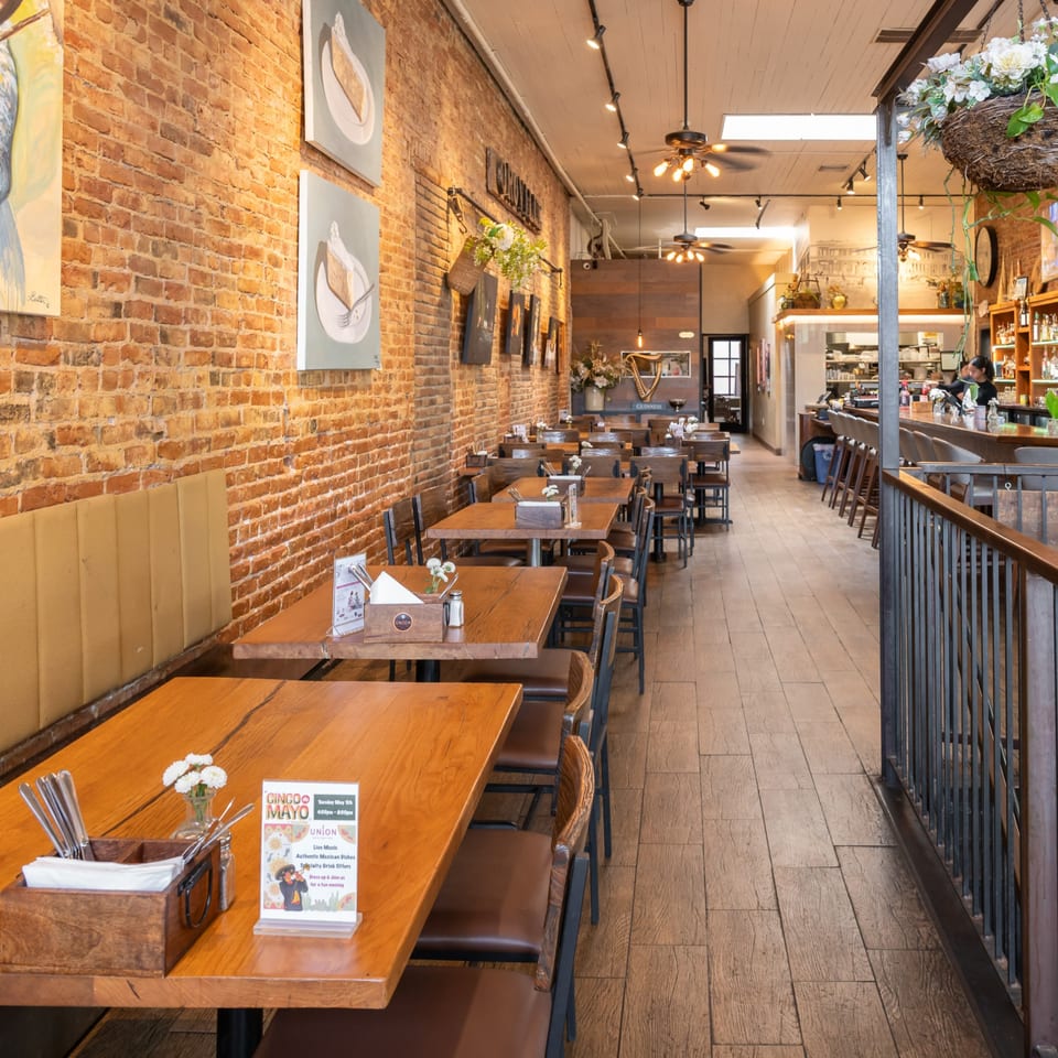 Long, narrow restaurant dining room with wooden tables and chairs, brick walls, and a bar area.