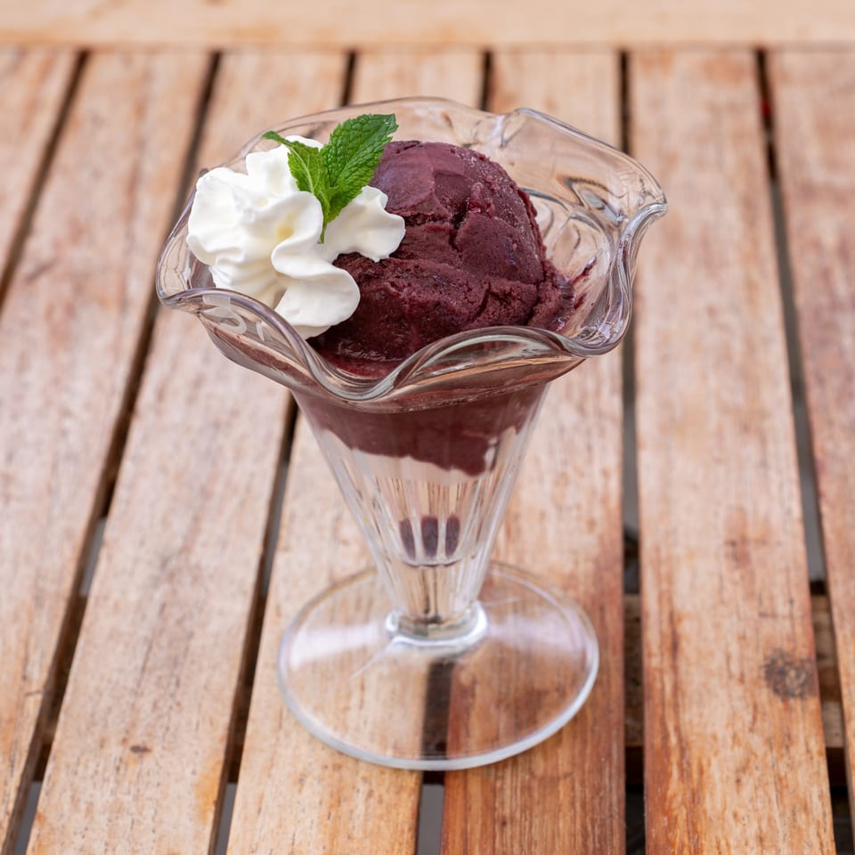 A scoop of dark purple ice cream with whipped cream and a mint leaf in a glass sundae dish.