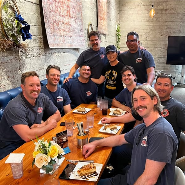 A group of firefighters and a woman in a Union shirt enjoy dessert at a long wooden table.