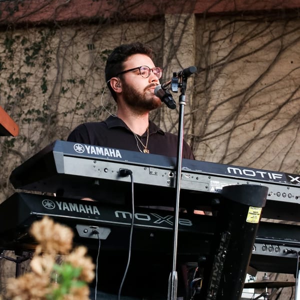 A man with glasses and a beard sings into a microphone while playing a Yamaha keyboard.