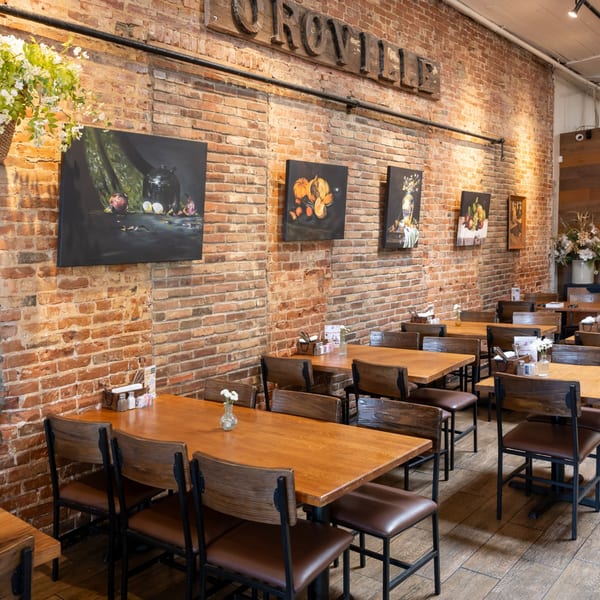 Restaurant interior with wooden tables and chairs, brick walls, and paintings, under a "OROVILLE" sign.