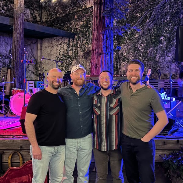 Four men smiling, standing in front of a stage with musical instruments and trees lit with blue and pink lights.
