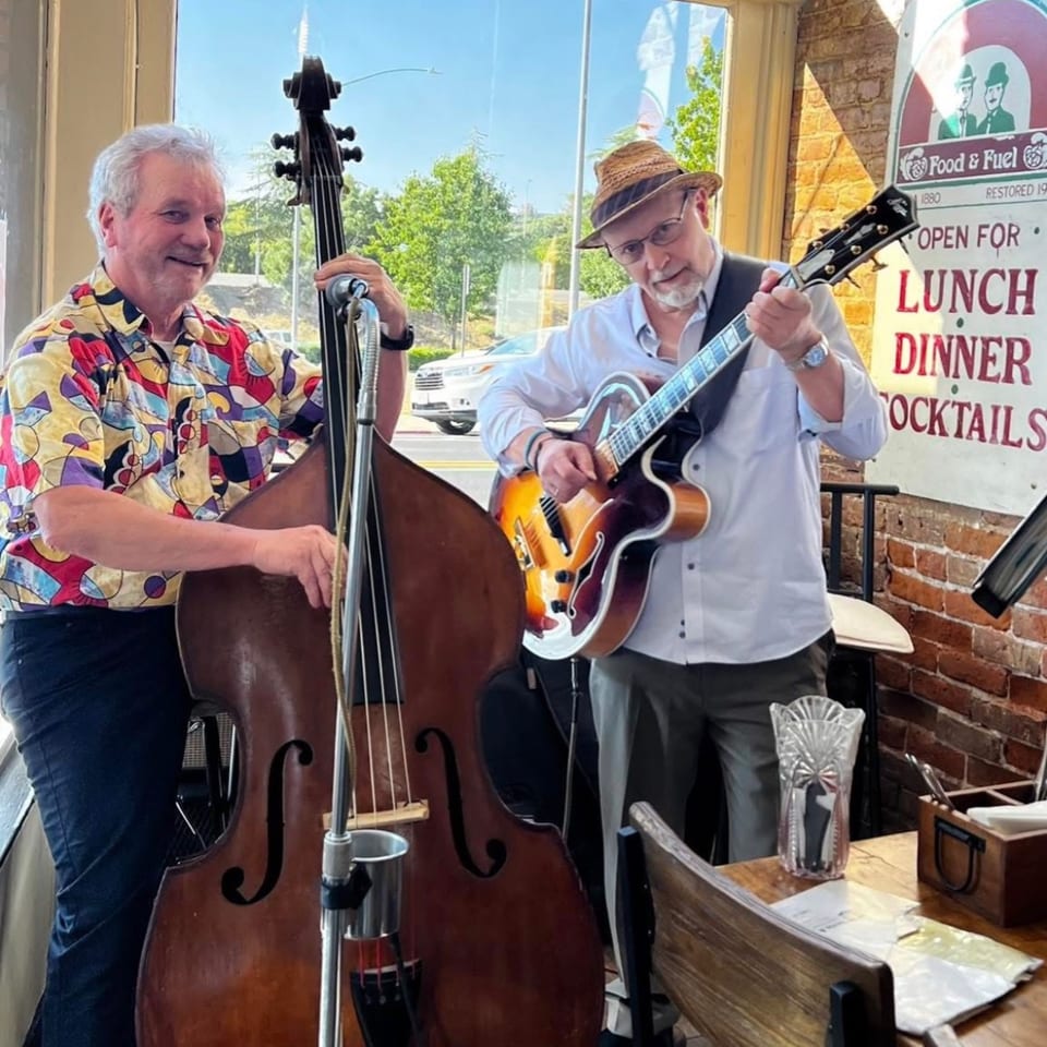 Two musicians, one playing an upright bass and the other a guitar, perform in a restaurant.