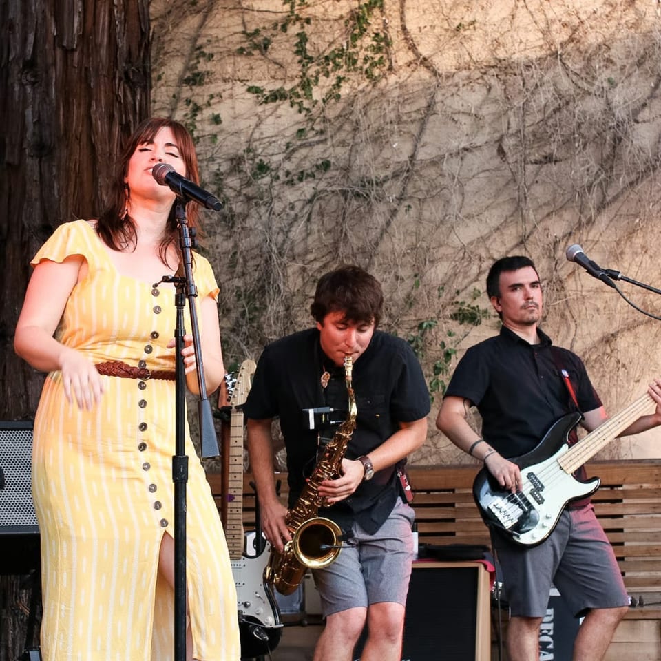 A female singer in a yellow dress performs with a saxophonist and bassist on an outdoor stage.