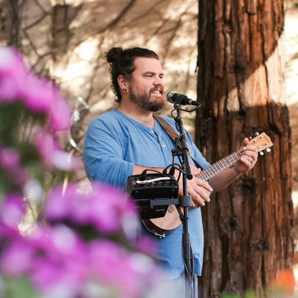 A man with a beard and man bun sings into a microphone while playing a ukulele outdoors.
