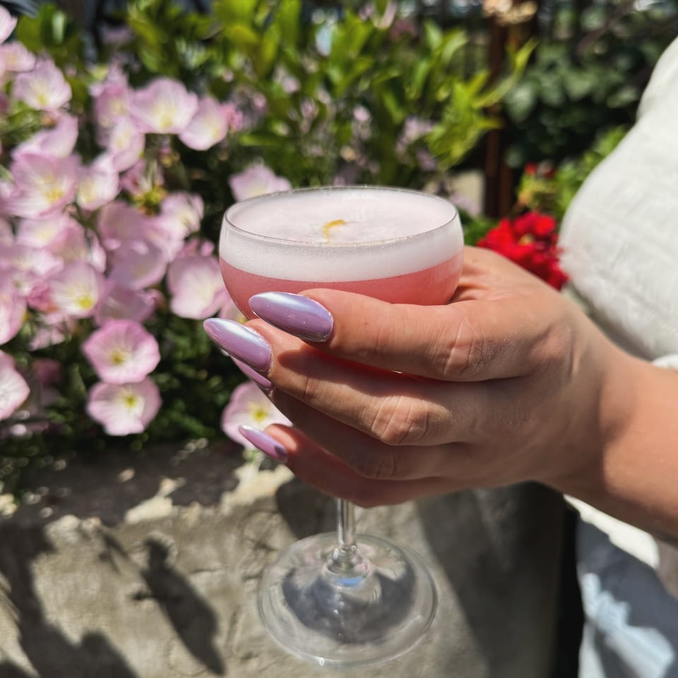 Hands with iridescent nails holding a pink foamy cocktail against a background of pink flowers.