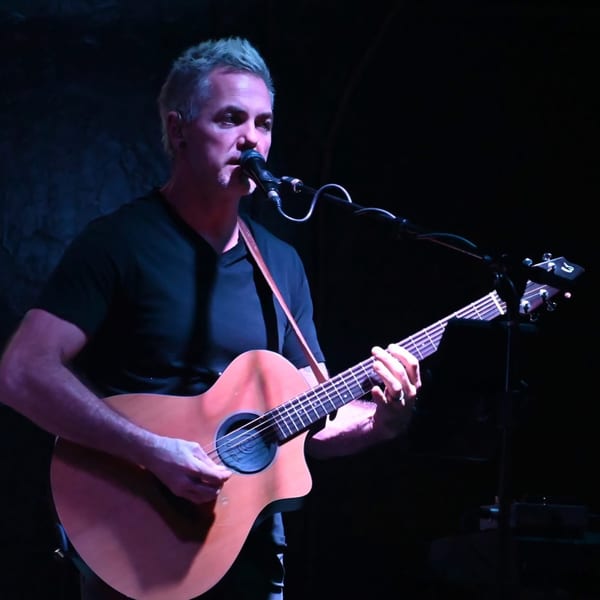 A male musician with gray hair sings into a microphone while playing an acoustic guitar on a dark stage.