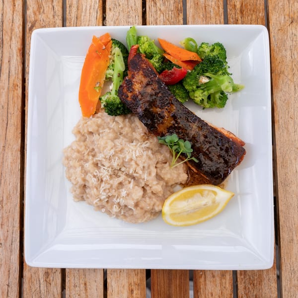 Glazed salmon with risotto, broccoli, carrots, and a lemon wedge on a white plate.