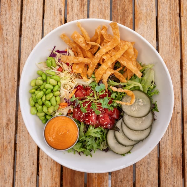 Overhead view of a colorful poke bowl with tuna, edamame, cucumbers, crispy wontons, and dressing.
