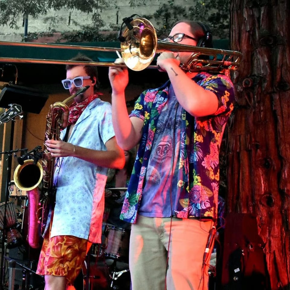 Two musicians in Hawaiian shirts play saxophone and trombone on a stage with red lighting.