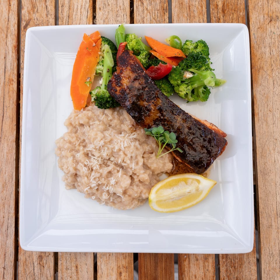 Glazed salmon with risotto, broccoli, carrots, and a lemon wedge on a white plate.