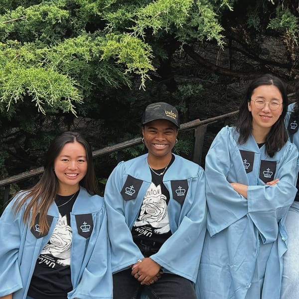 Three smiling students in light blue graduation gowns with Union branding, standing outdoors.