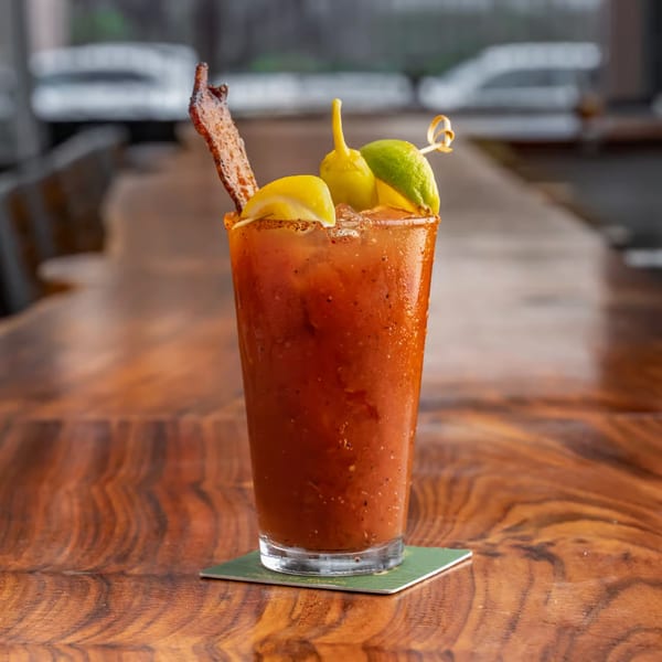 A Bloody Mary cocktail with bacon, lemon, lime, and pepper garnishes on a wooden table.