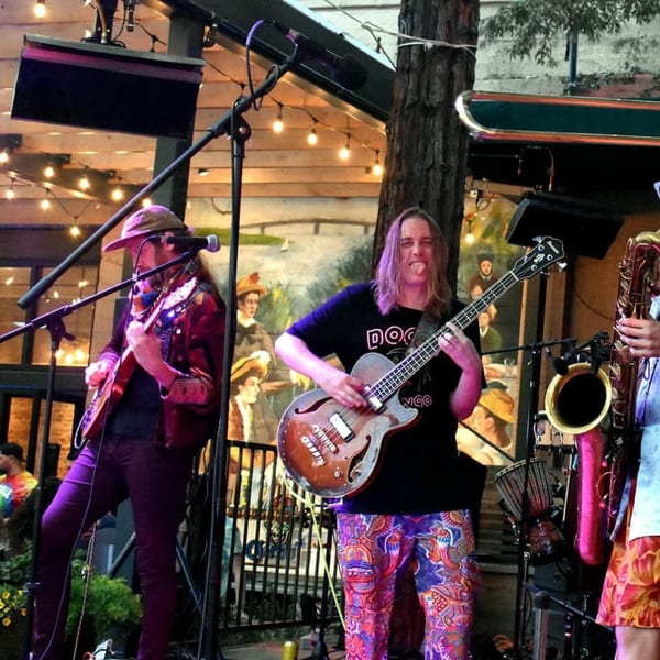 A band performs live on an outdoor stage with string lights and a mural in the background.