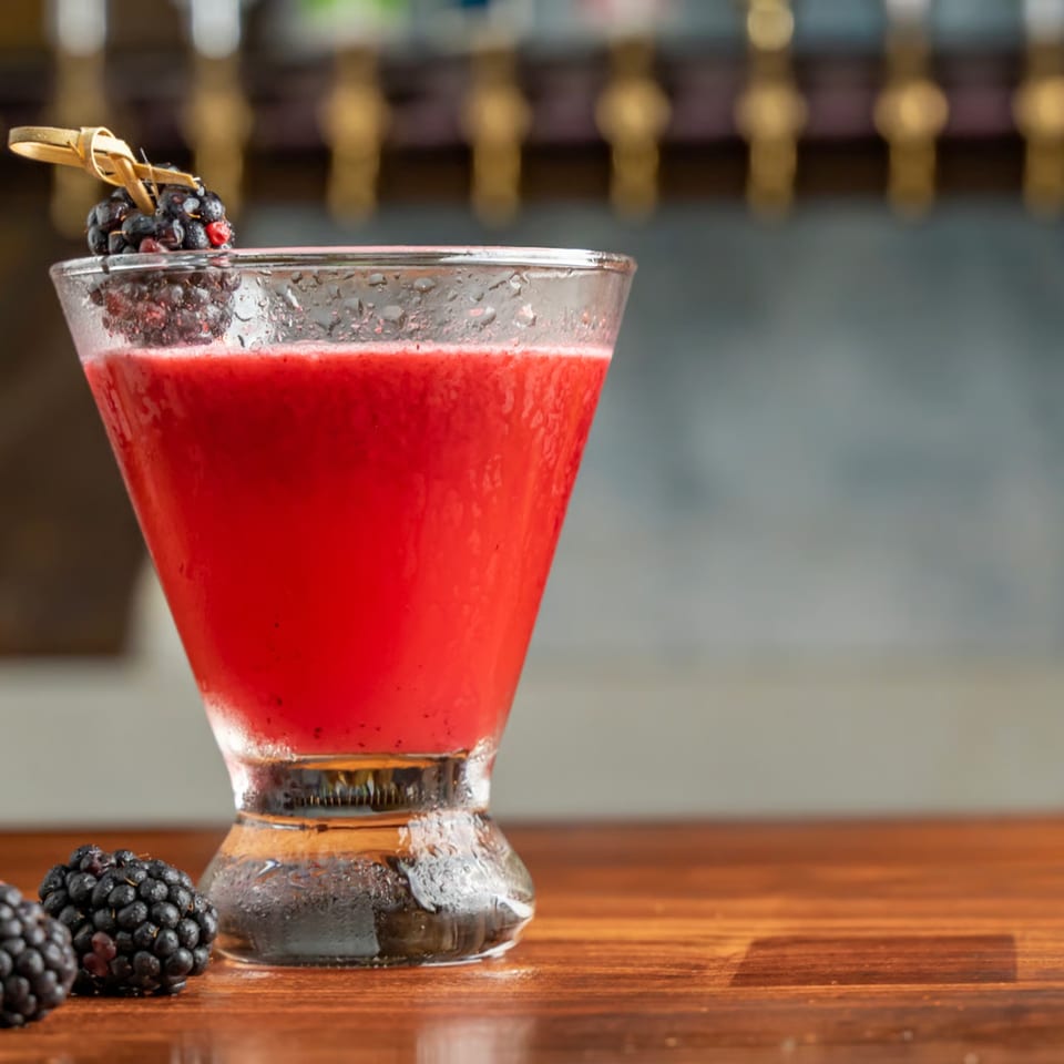 A vibrant red cocktail in a martini glass, garnished with fresh blackberries, on a wooden bar.
