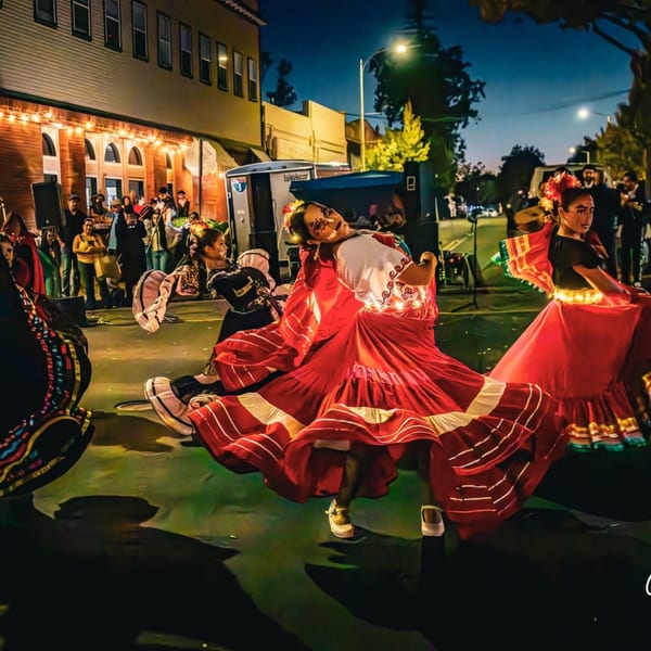 Dancers in vibrant traditional dresses perform on a street at night, with a crowd and buildings in the background.
