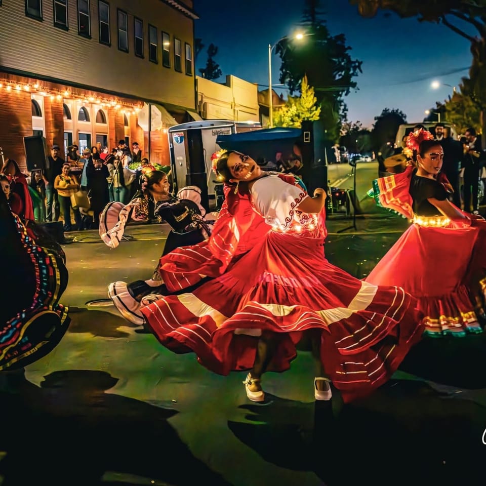 Dancers in vibrant traditional dresses perform on a street at night, with a crowd and buildings in the background.