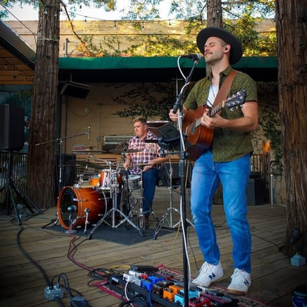 A musician in a hat plays guitar and sings, with a drummer behind him on an outdoor stage.
