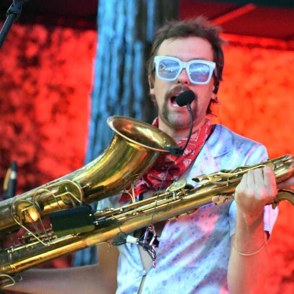A man with a mustache and white sunglasses plays a saxophone with a microphone attached.