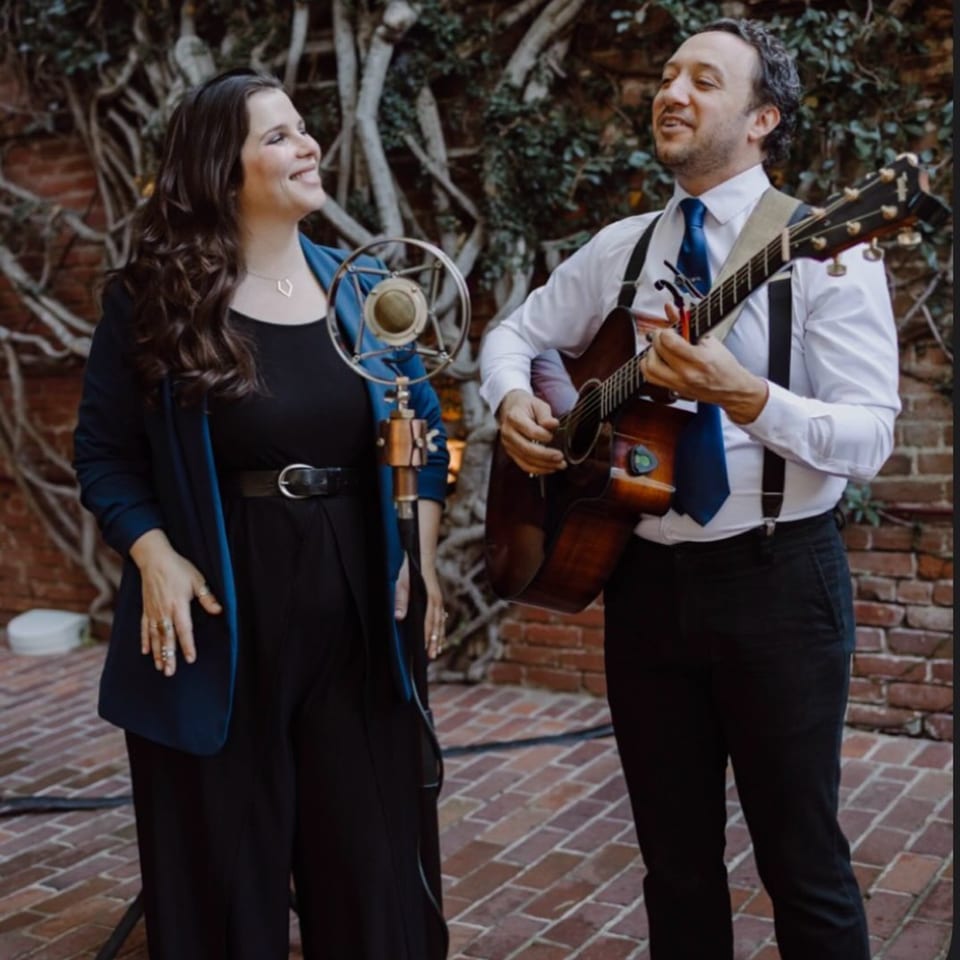 A woman singing into a vintage microphone next to a man playing an acoustic guitar outdoors.