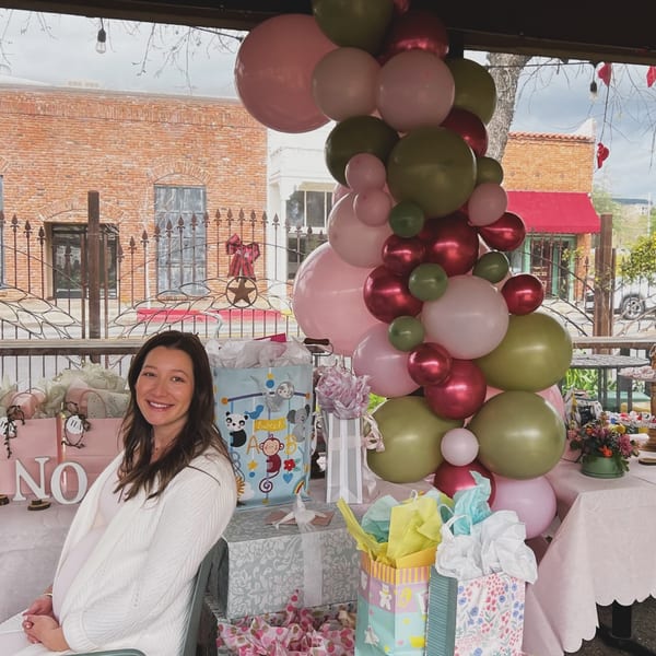 A smiling pregnant woman sits at a table with gifts and balloons on an outdoor patio.