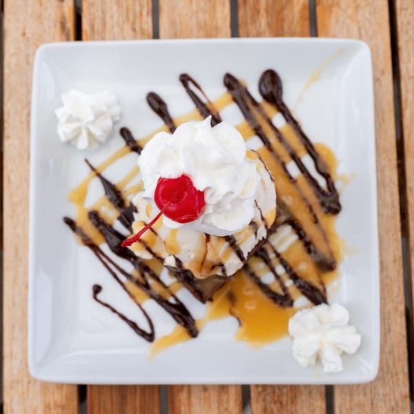 Dessert on a white plate with ice cream, whipped cream, a cherry, caramel, and chocolate drizzle.