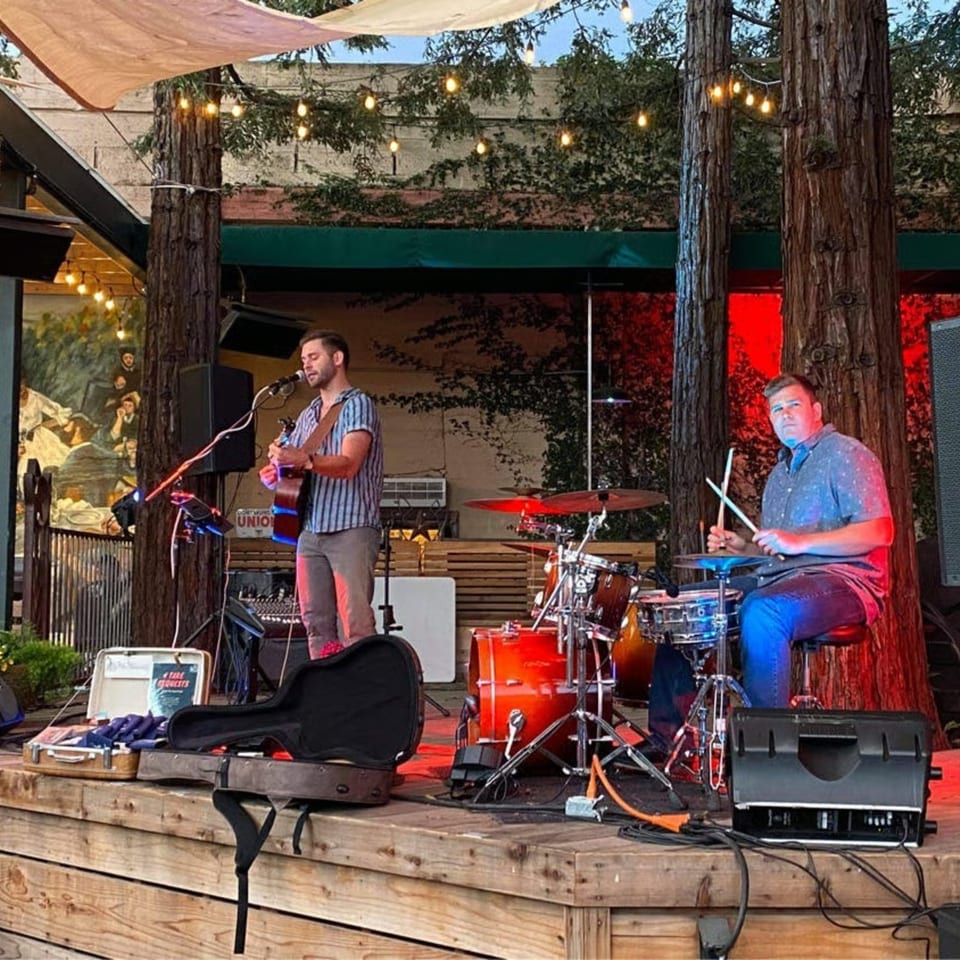 Two musicians perform live on an outdoor stage at Union, with string lights and trees.