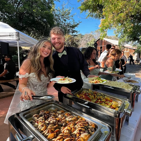 Smiling couple serving themselves American cuisine from a buffet at an outdoor event.