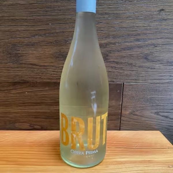 A bottle of Opera Prima Brut sparkling wine with a light blue cap on a wooden surface.