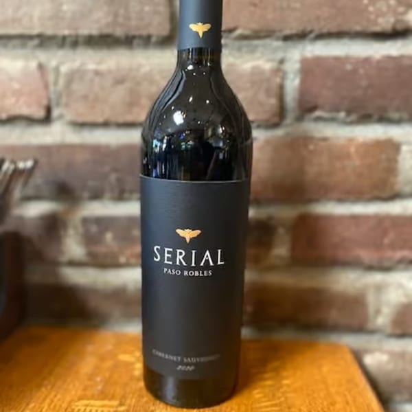 A bottle of Serial Paso Robles Cabernet Sauvignon wine stands on a wooden surface against a brick wall.