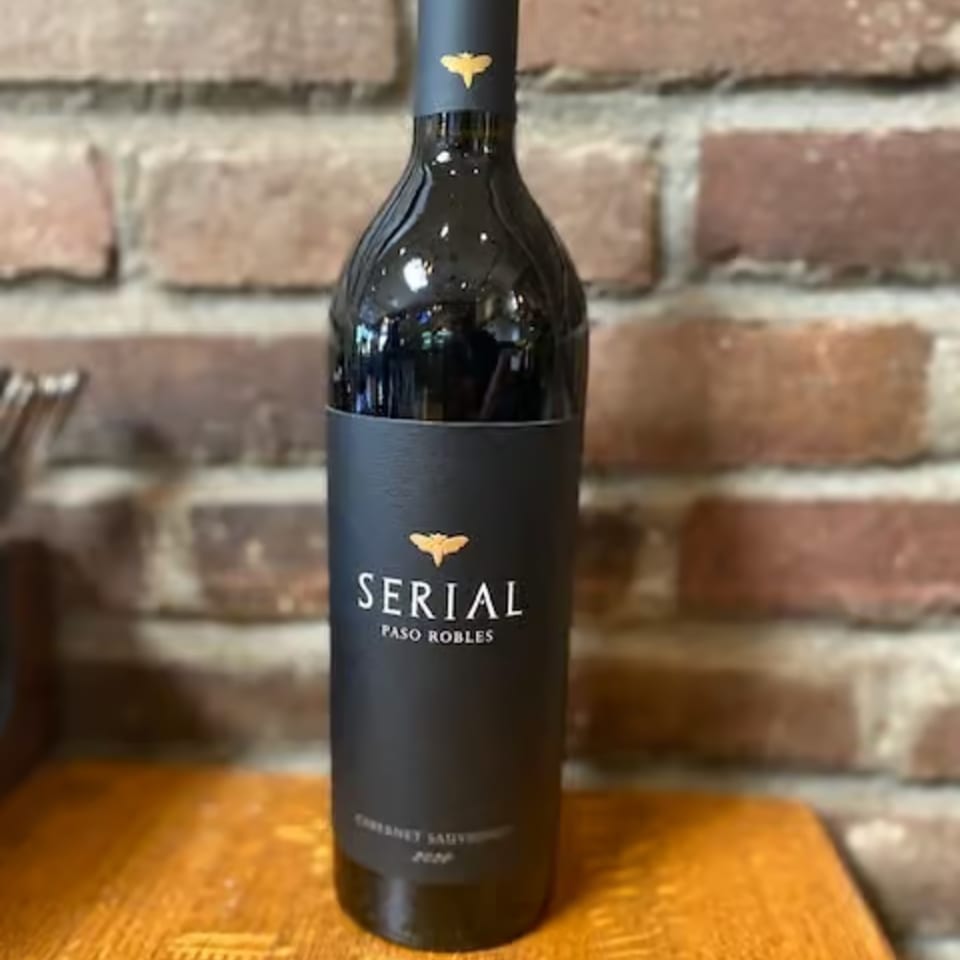 A bottle of Serial Paso Robles Cabernet Sauvignon wine stands on a wooden surface against a brick wall.