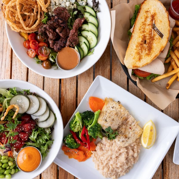 Overhead view of a wooden table with three dishes: steak salad, poke bowl, and fish with risotto.