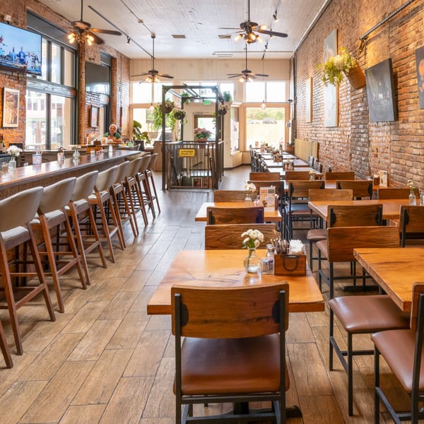 Spacious restaurant interior with a long bar, dining tables, brick walls, and large windows.