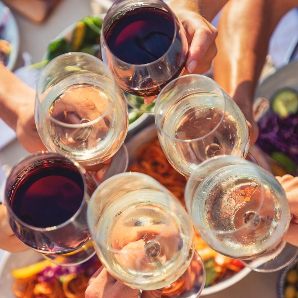 Hands holding six wine glasses, some red and some white, toasting over a meal.